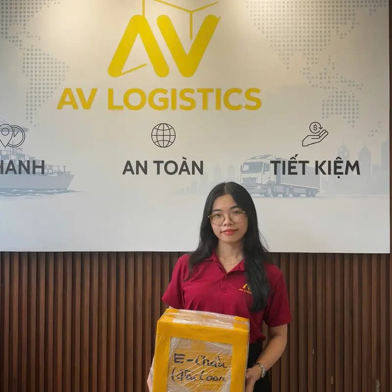 Nhân viên cầm thùng hàng đã đóng gói trước bảng hiệu AV Logistics